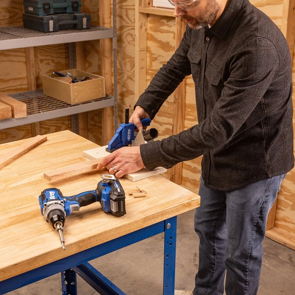A cordless drill with a battery pack sits on a woodworking workbench, with woodworking clamps nearby and an operator wearing safety glasses working beside it.